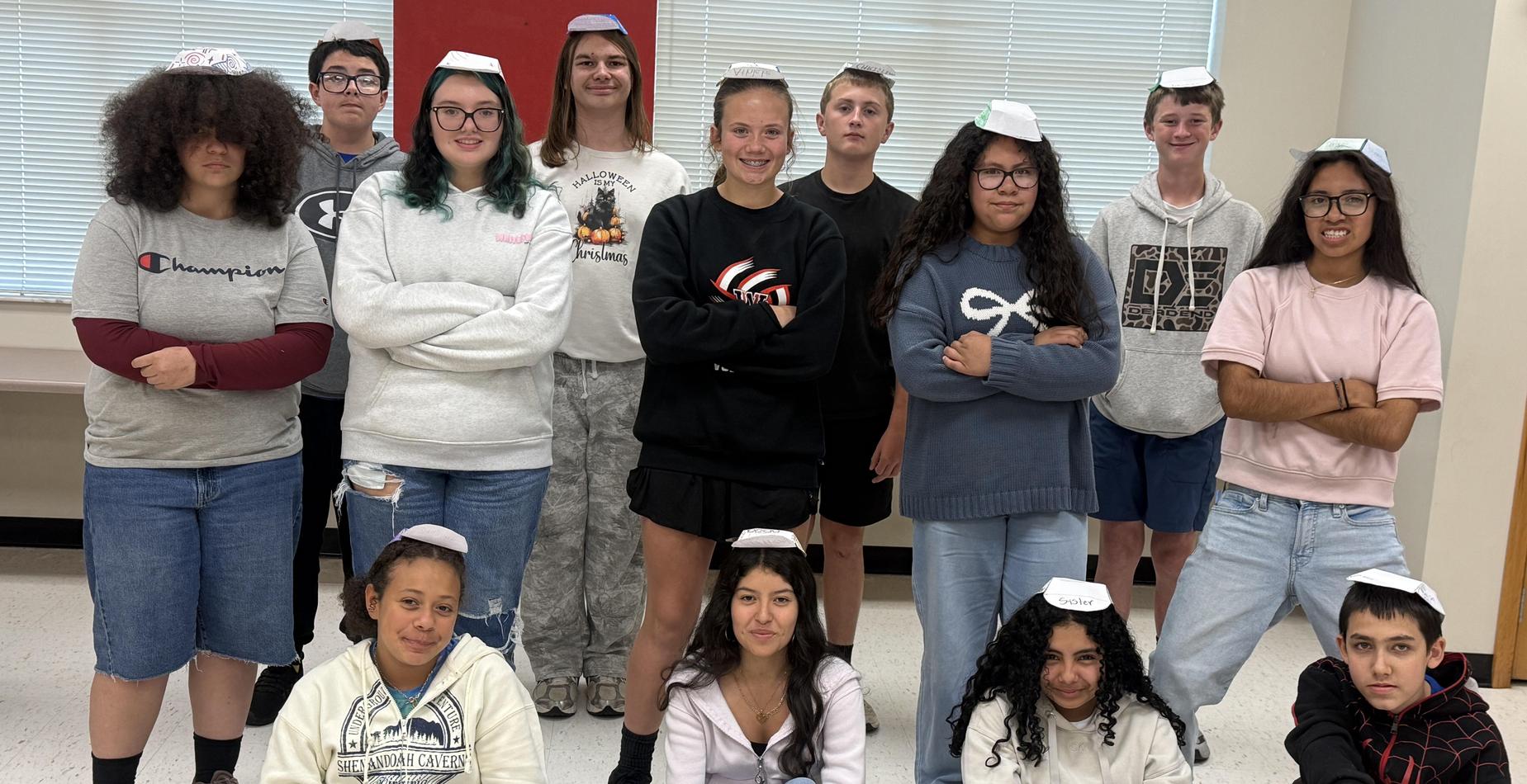 Group of teens in a classroom wearing paper hats and posing together.