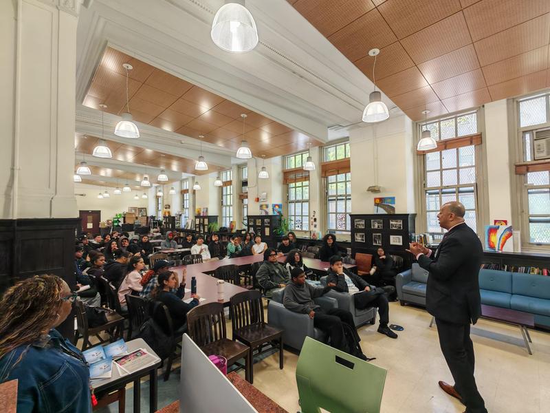 Audience listening attentively in a large room with seating and artwork.