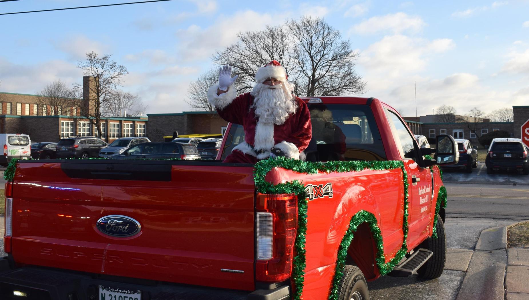 Santa Claus waving from the back of a red truck decorated with garland.