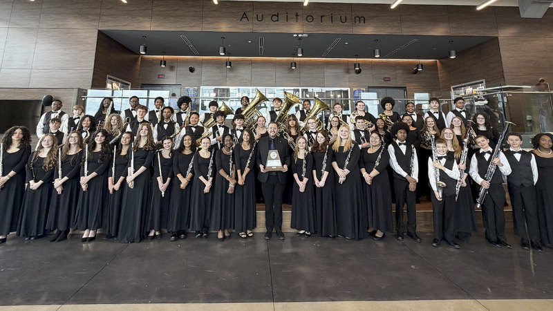A band in concert uniform smiling with a trophy in an auditorium lobby