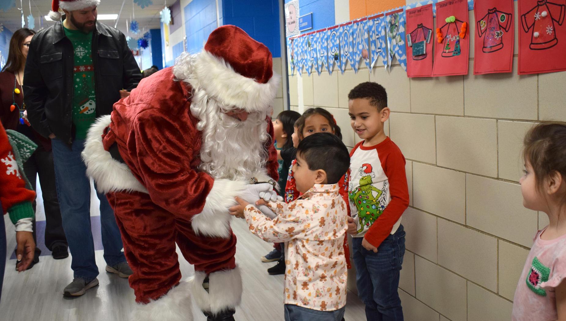 Santa Claus greeting a young boy while children watch excitedly in a school hallway.