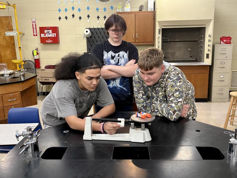 Three students hovering around a balance measuring glue sticks' weight