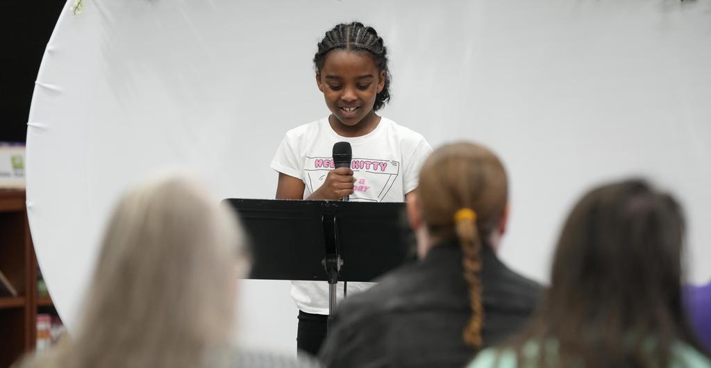 A student speaks into a microphone to a group of adults.