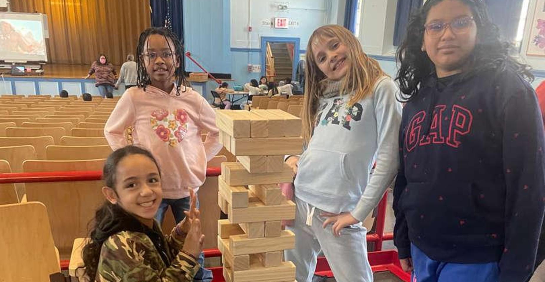 Four children posing beside a tall stack of wooden blocks in a gym setting.