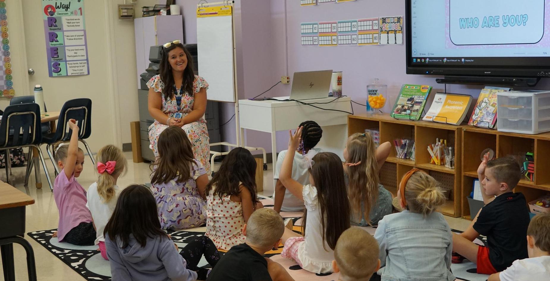 Students sitting on the floor raising hands during a lesson with a teacher.