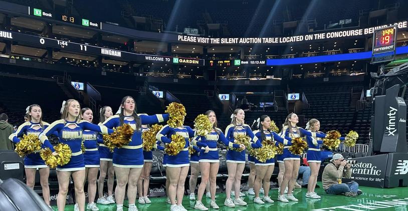 Cheerleaders performing in a sports arena with a scoreboard visible in the background.