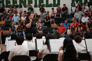 Female conductor leads an orchestra and students dressed in white shirts await their cue