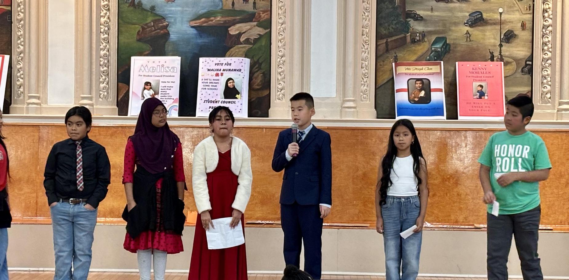 Children presenting in formal attire at a school event with posters in the background.