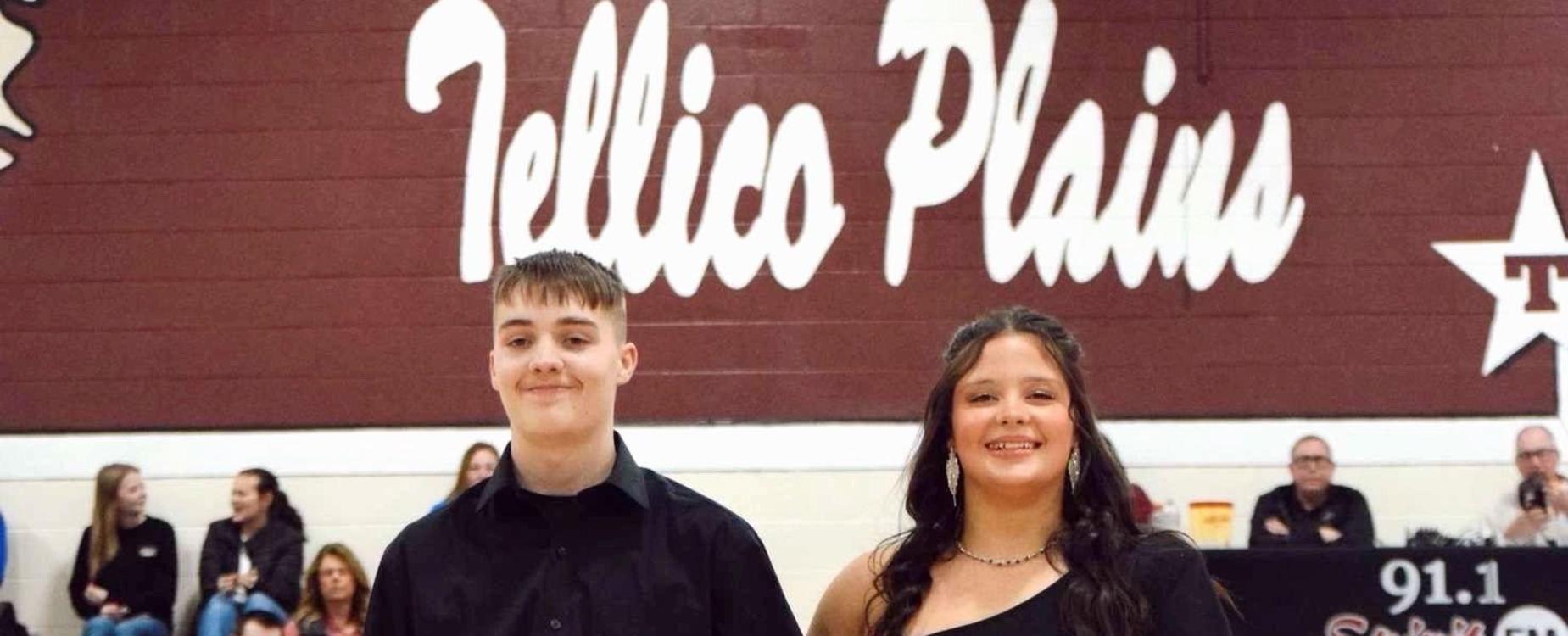 A couple walking together on a gym stage, one in a formal outfit and the other in a dress.
