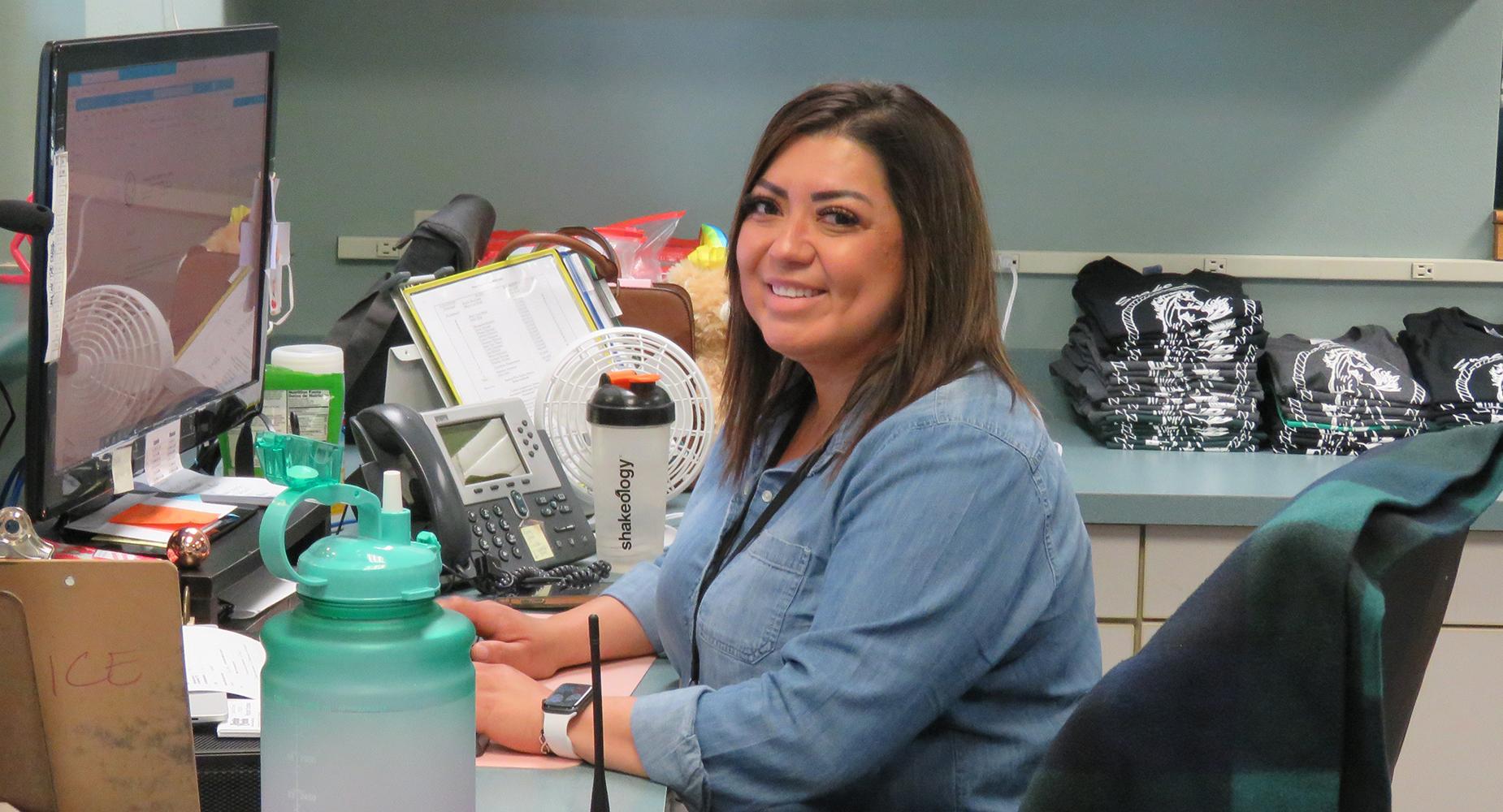 School secretary sitting at her front office desk faces the camera and smiles