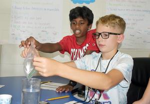Two boys working together on a science experiment, handling a bag and water at a table.