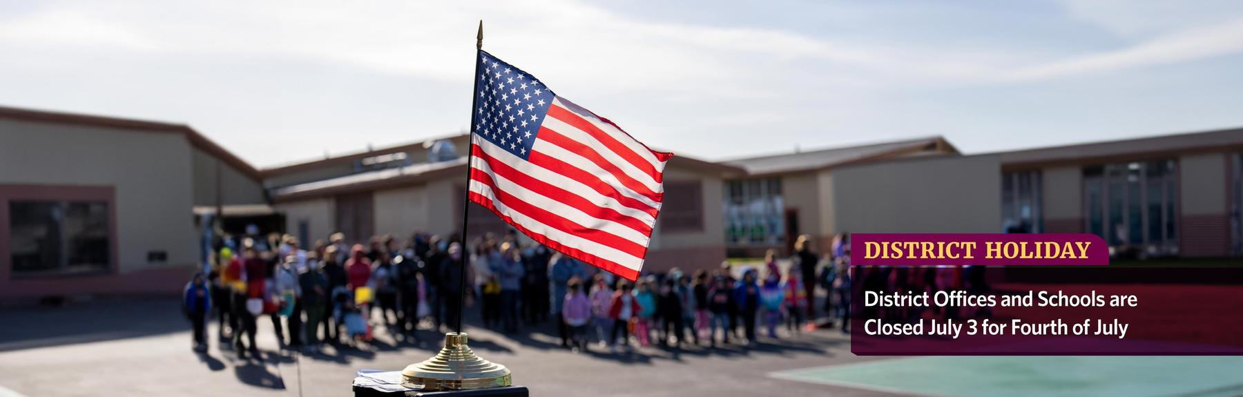 An American flag on a pedestal with a school playground assembly in the background, and the words: District Holiday: District Offices and Schools Closed July 3 for Fourth of July.