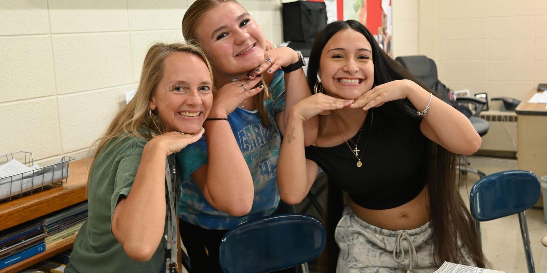 Three smiling women posing playfully with their hands on their chins in a classroom.