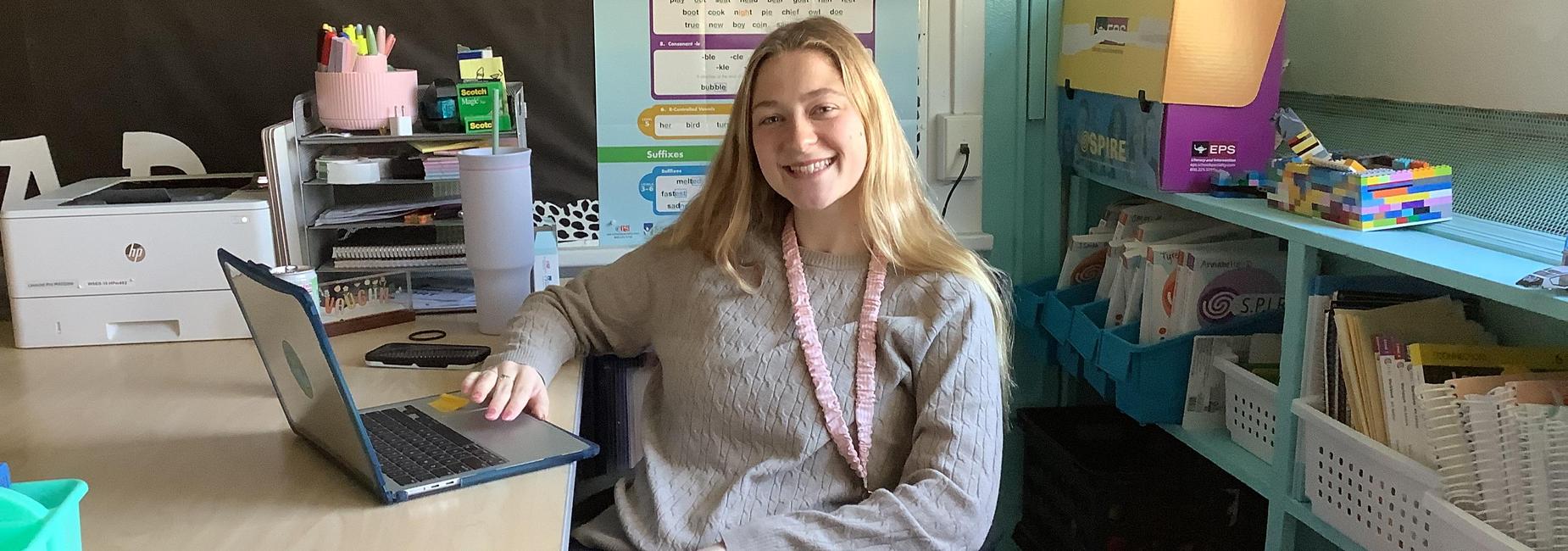 Smiling woman sitting at a desk with a laptop in a classroom setting.