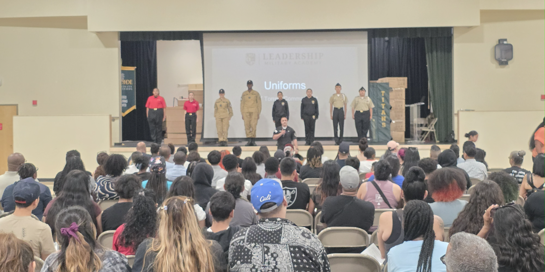 A crowd gathered in an auditorium watching a presentation featuring speakers in uniforms.