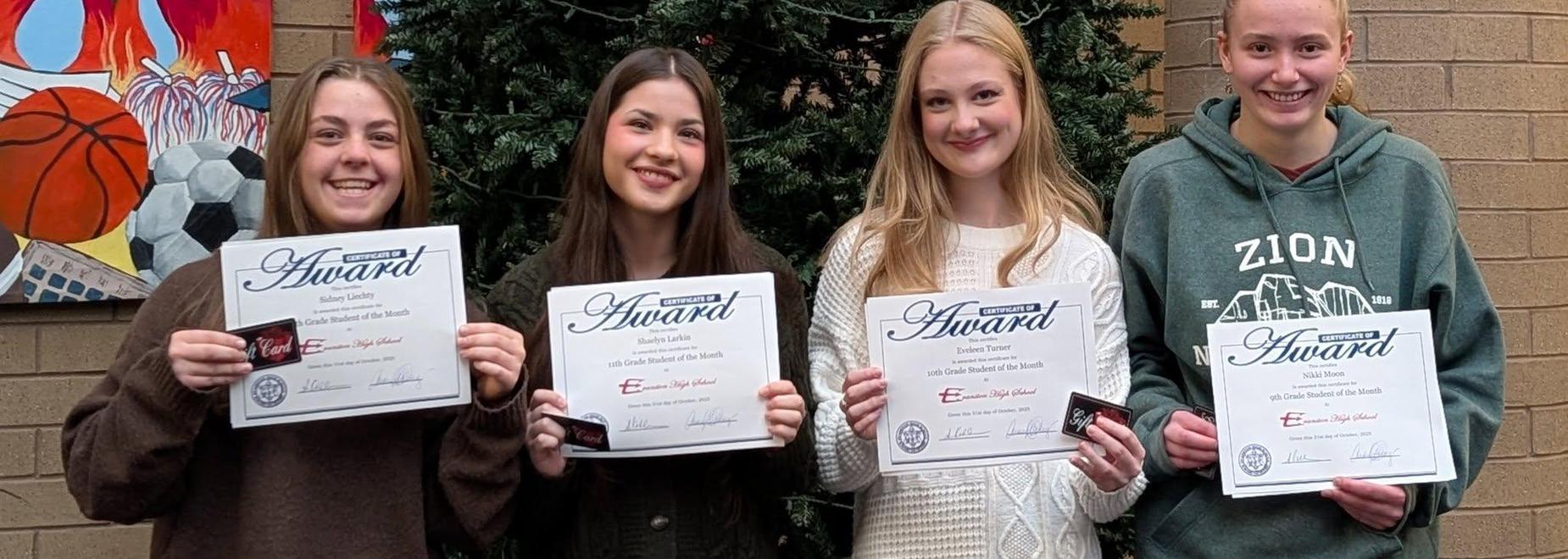 Four students holding awards and smiling in front of a decorated wall.