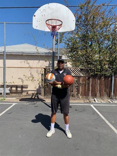 Man holding a basketball and a volleyball on a court under a hoop.