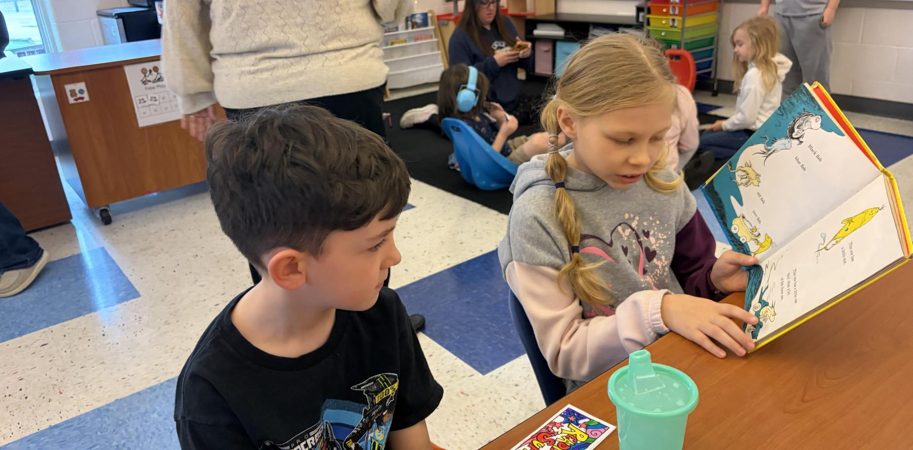 A girl reads to a boy at a classroom table.