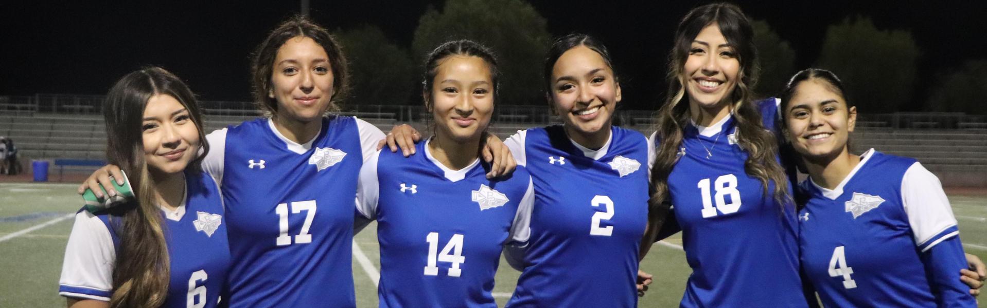 Six female soccer players in blue jerseys smiling together on a field.