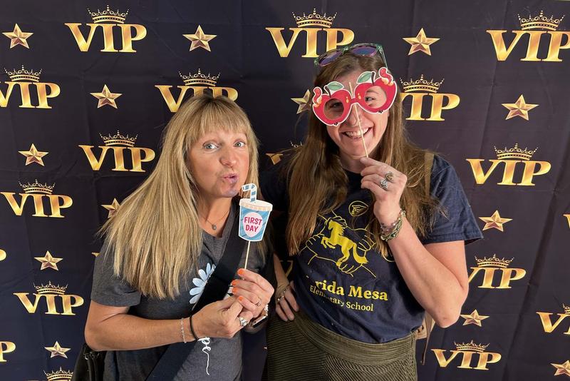 Two women pose in front of a black and gold “VIP” backdrop, holding playful props including a “First Day” sign and apple-shaped glasses.