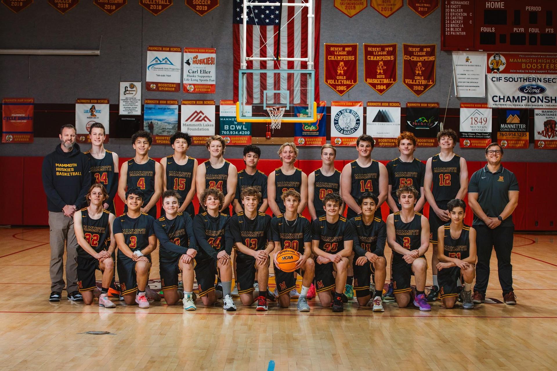 Basketball team posed on court with banners and a hoop in the background.