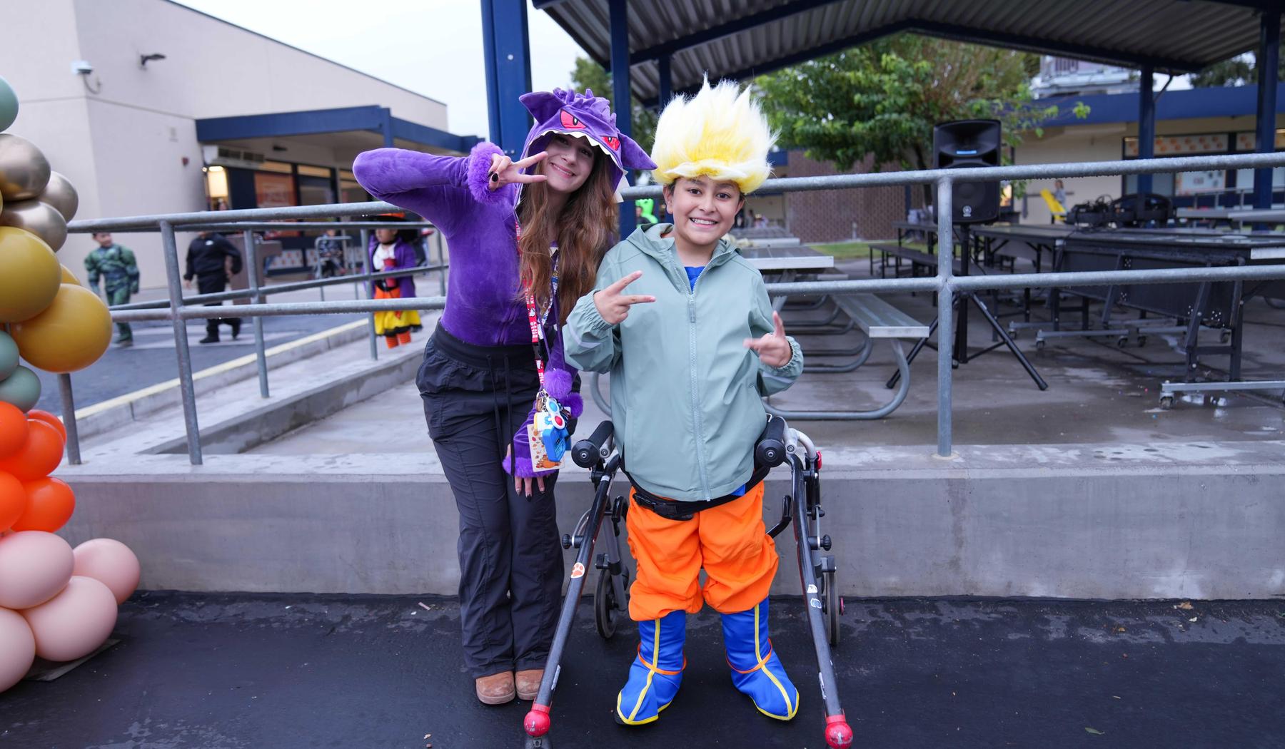 A girl in a purple costume poses with a boy in a colorful outfit and wheelchair.