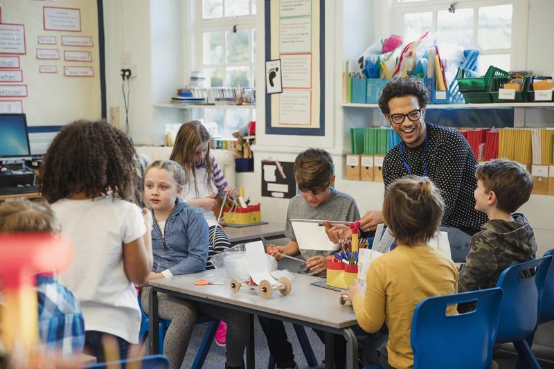 A smiling male teacher wearing glasses interacts with a group of young students working on a hands-on crafting project at a classroom table.