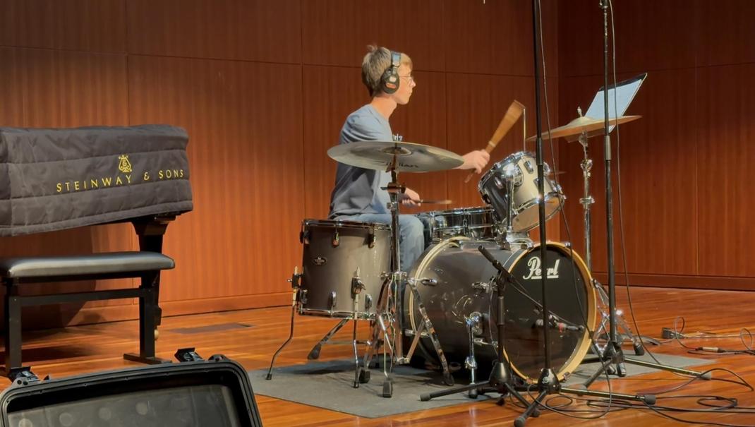 A drummer plays a drum kit on stage beside a grand piano in a concert hall.