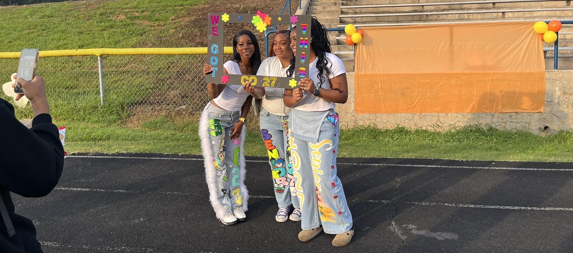 Three young women pose with a colorful frame celebrating their graduation year.