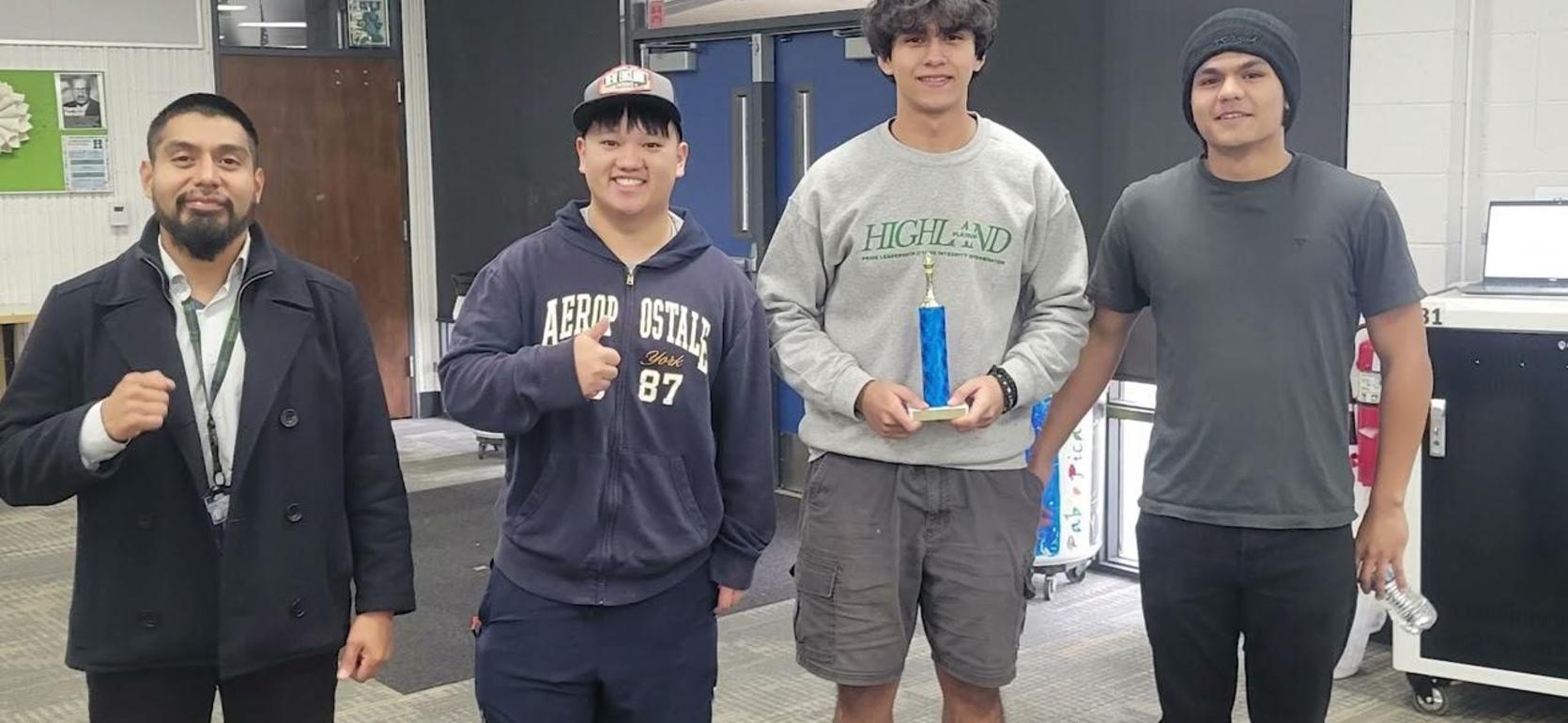 Three young men stand with a trophy in a classroom alongside a teacher.