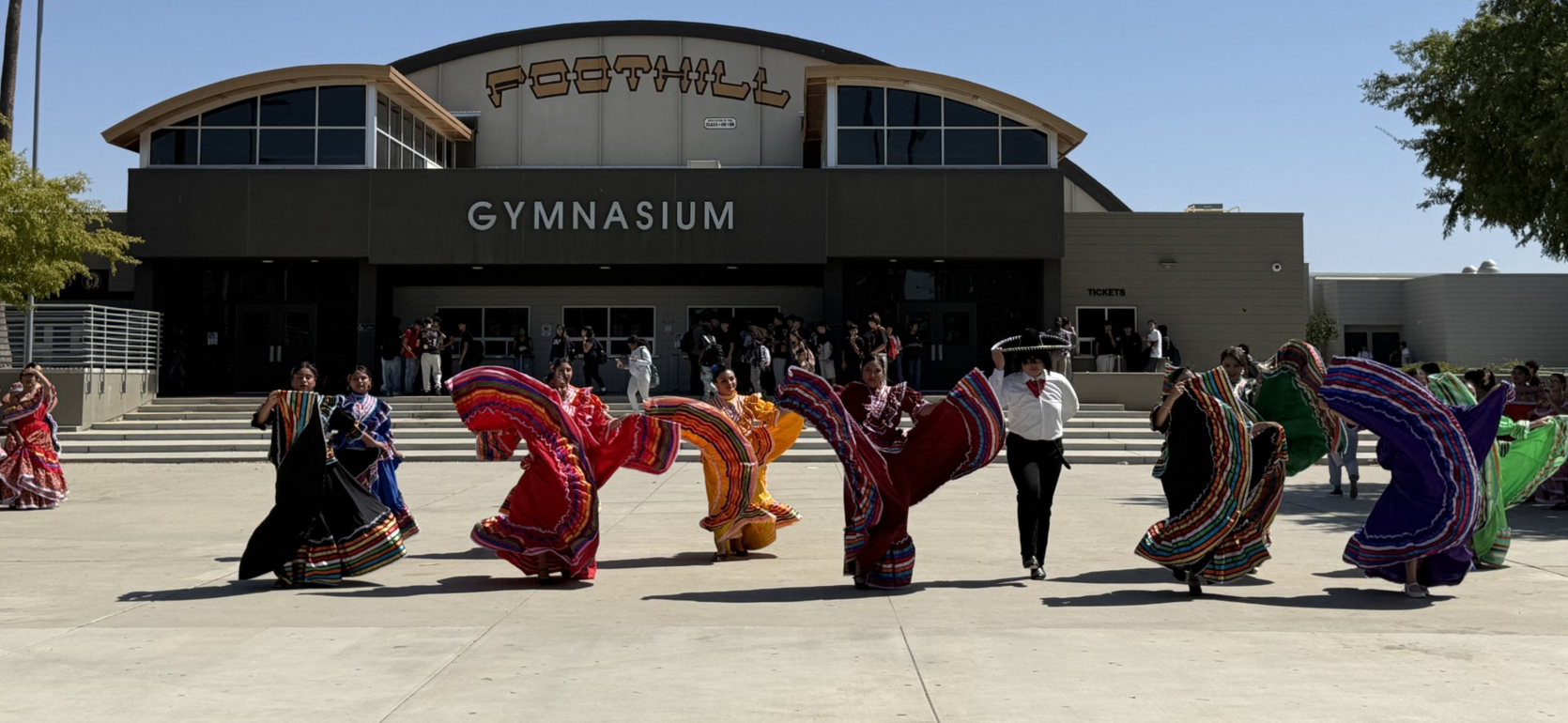 Dancers in colorful dresses perform near a gymnasium under a clear blue sky.