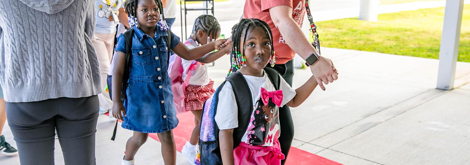 Young girls with colorful hairstyles walking hand-in-hand with an adult through school entrance.