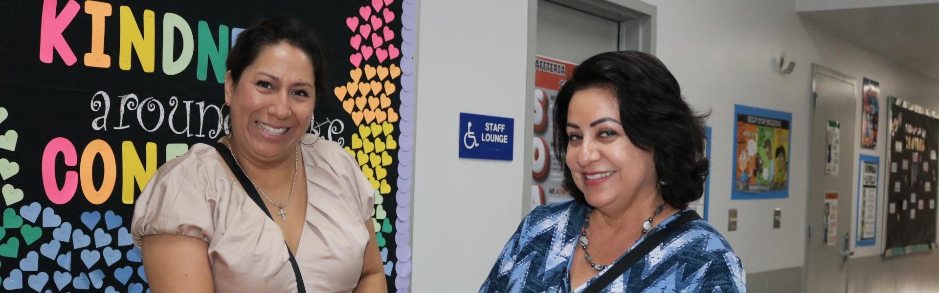 Two women smiling together in a school hallway with colorful bulletin boards.