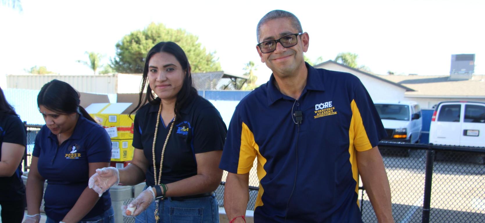 Two individuals serving food at an outdoor event with a table covered in food.