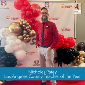 Nicholas Patey, a bearded man with glasses, poses in front of the Los Angeles County Office of Education Teacher of the Year banner with red and gold balloons.