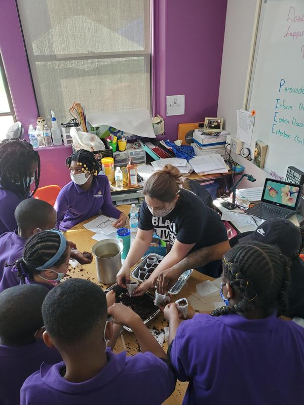 Scholars and their teacher gather around a table while the teacher demonstrates a planting technique.