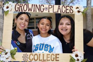 Three students smile behind a decorated “Growing Places” photo frame featuring flowers and a “#College” sign.