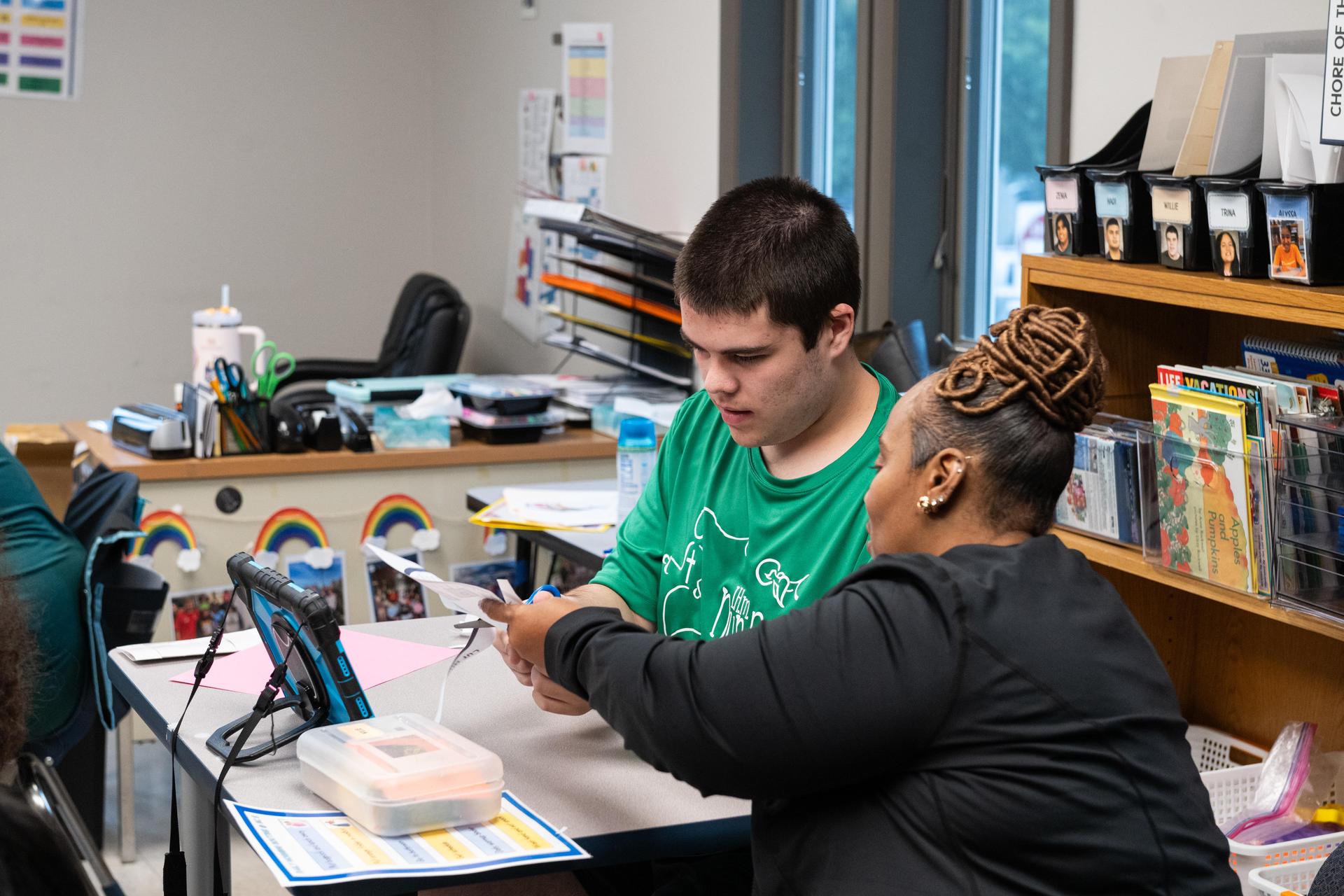 Student in green looking at assignment with teacher guiding.