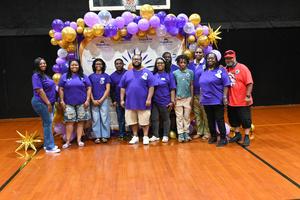 Group of attendees in purple shirts posing in front of a balloon backdrop.