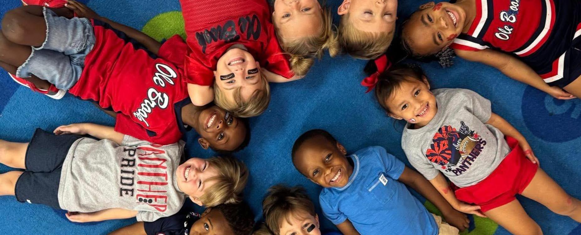 Group of ten children lying together on a colorful carpet, smiling and facing the camera.