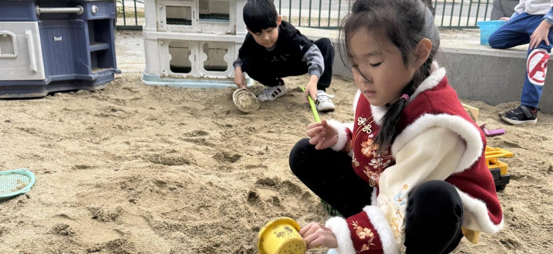 Child playing in sand, using a yellow cup in an outdoor sandbox.