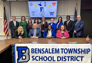 Image of 13 woman and men 4 sitting at the meeting room table and 9 standing behind them with text that says Bensalem Township School District