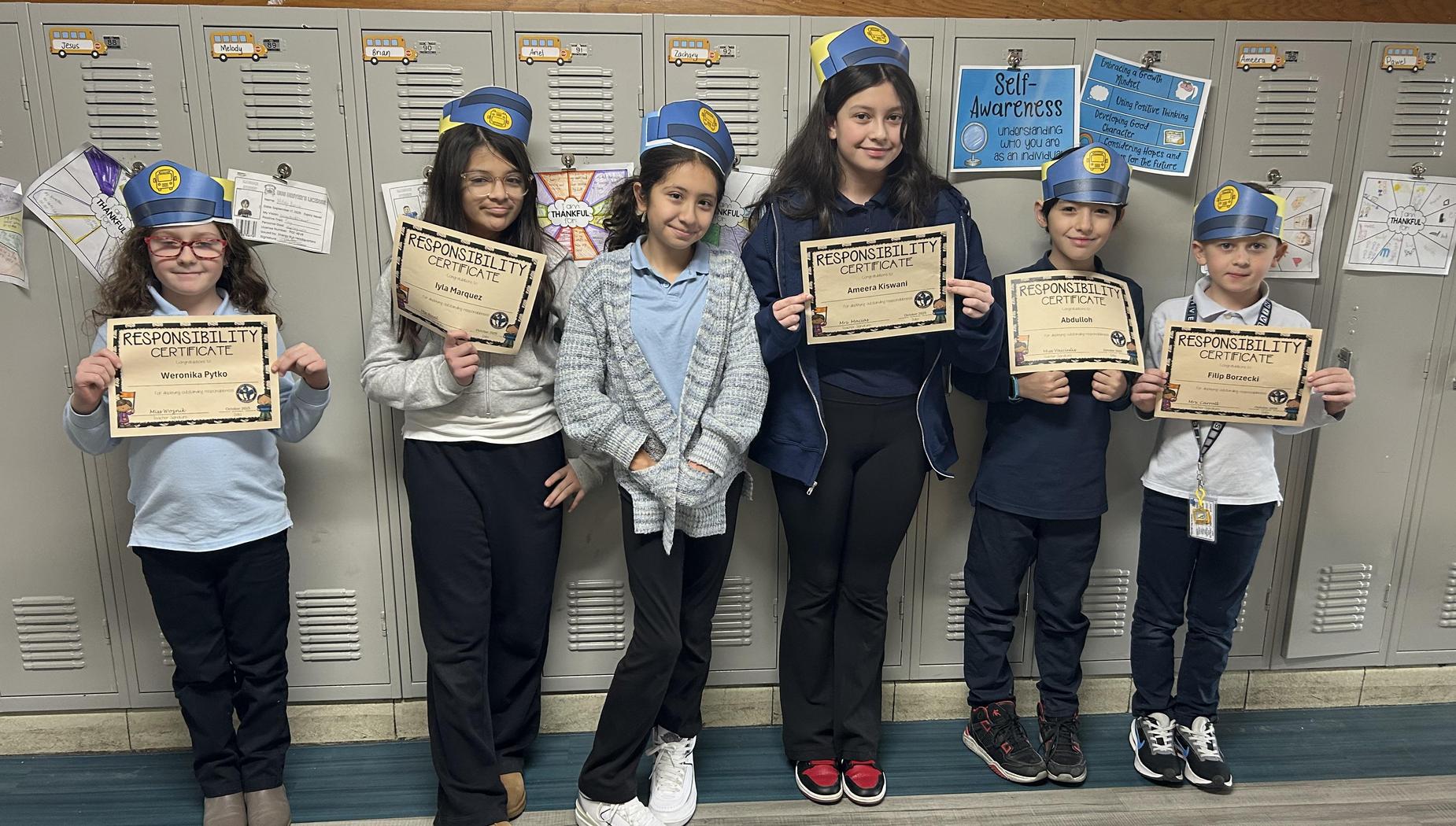 Six children wearing blue hats stand in front of lockers holding responsibility certificates.