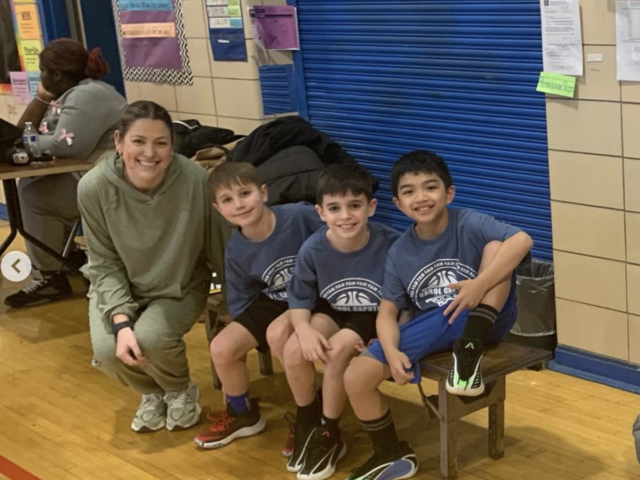 A coach sitting with three young boys, all wearing basketball t-shirts on a gym bench.