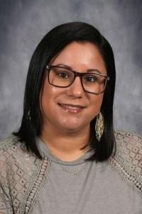 Smiling woman with straight hair wearing glasses and earrings, with a gray sweater.