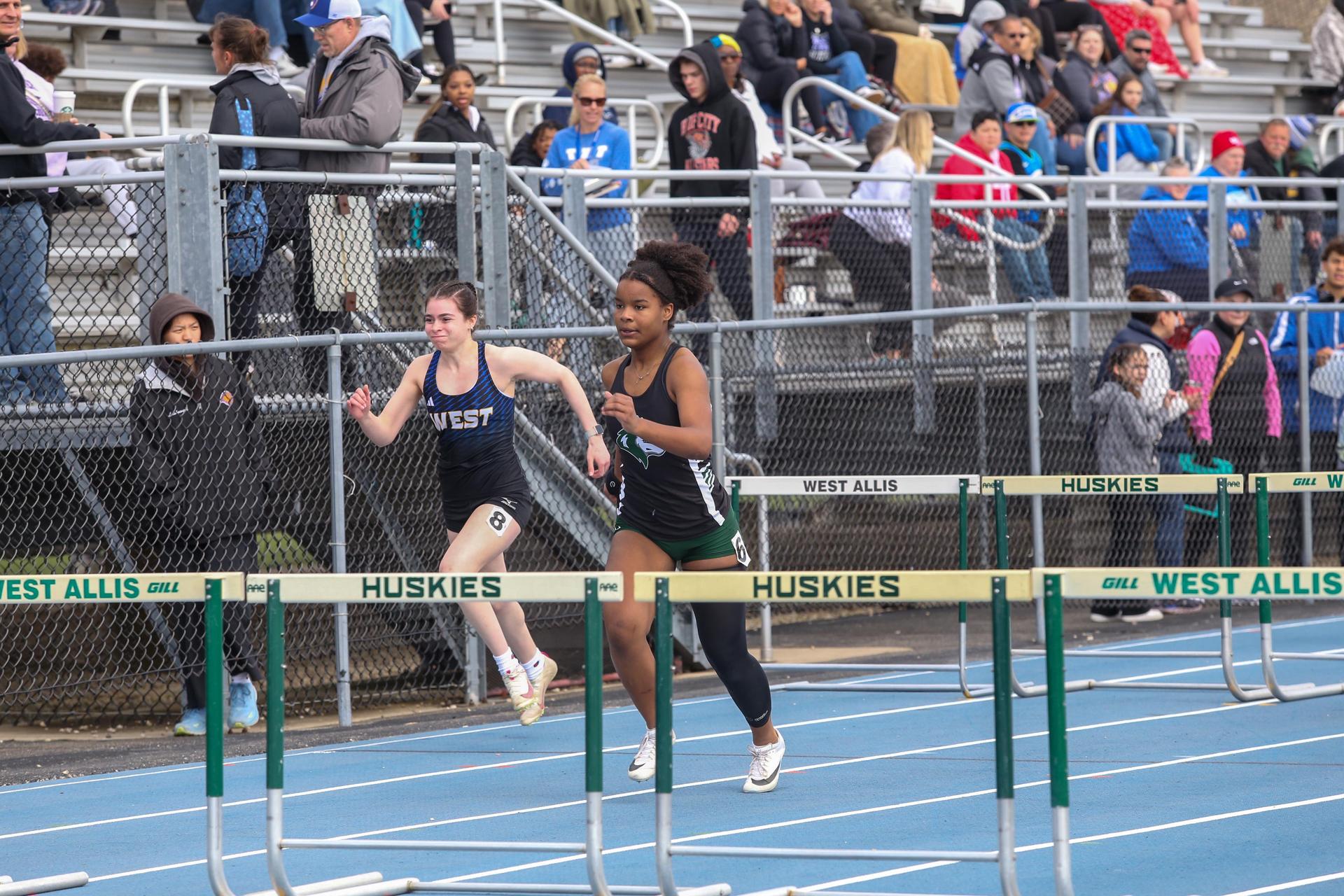 Two female athletes competing in a hurdle race on a track, with spectators in the background.