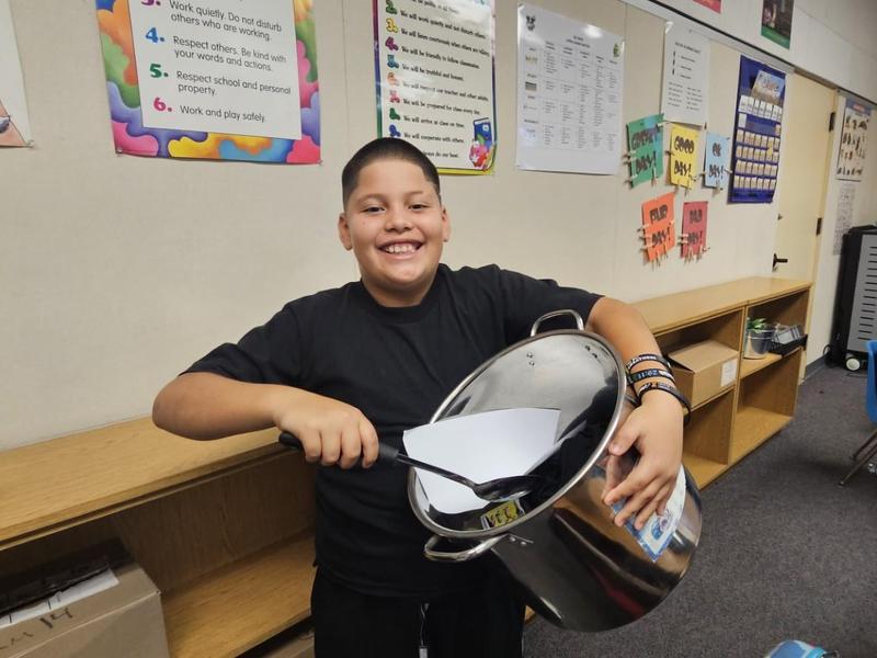 Smiling boy holding a large pot filled with papers in a classroom.
