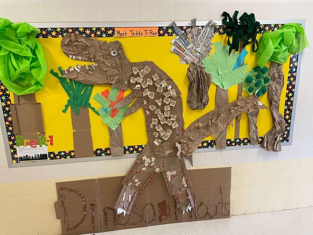 A bulletin board display featuring a T-Rex made of textured paper and green decorations.