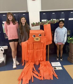 Three children stand beside a large orange gorilla made of paper strips.