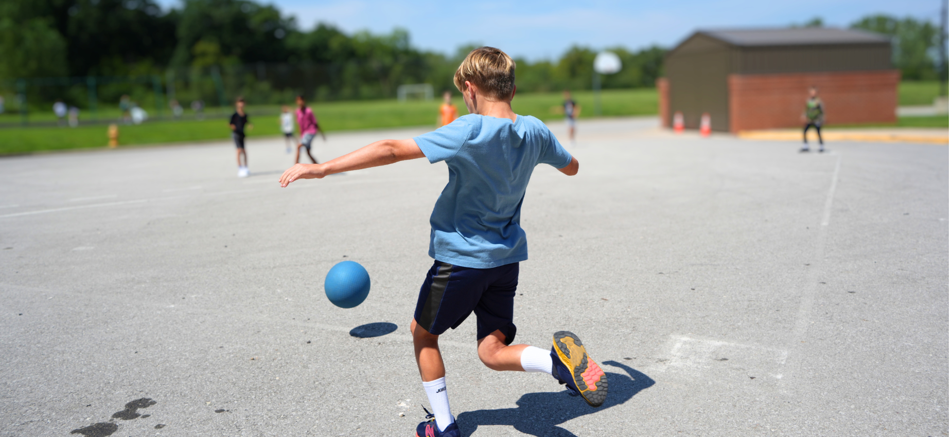 Boy kicking a blue ball on a playground with other children in the background.